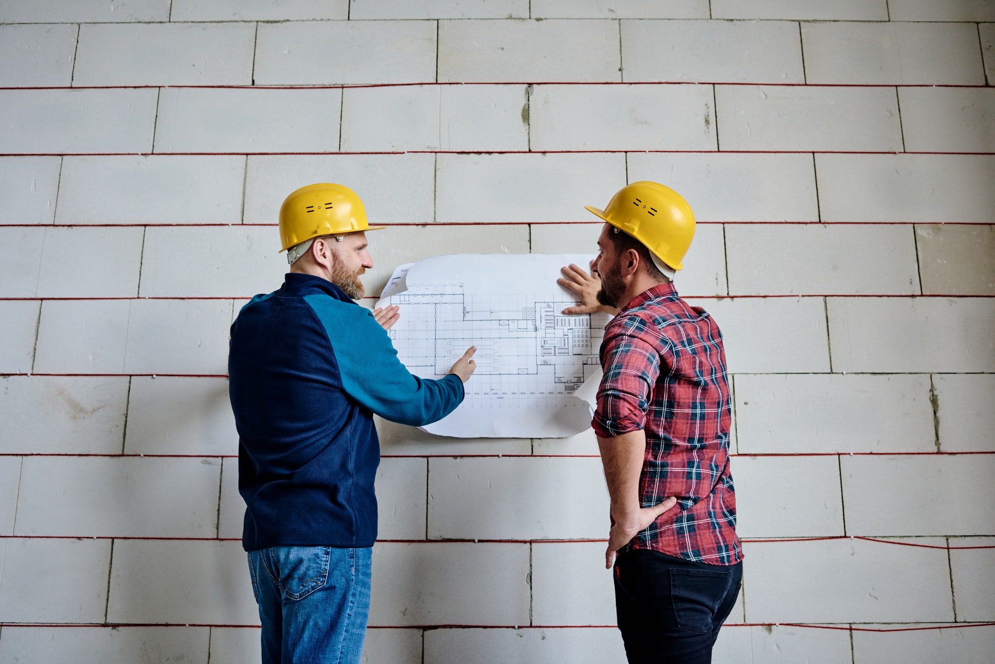 Two successful builders in protective helmets discussing blueprint by wall